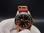 The Vintner SJT Original wristwatch with a gold-tone case, dark brown dial, red and blue bezel, and a red fabric strap, held in a white gloved hand against a dark background.