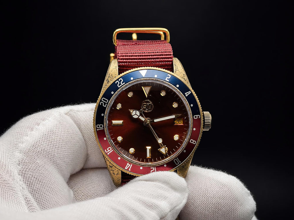 The Vintner SJT Original wristwatch with a gold-tone case, dark brown dial, red and blue bezel, and red fabric strap, held by a white gloved hand against a black background.