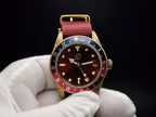 The Vintner SJT Original wristwatch with a gold-tone case, dark brown dial, red and blue bezel, and red fabric strap, held by a white gloved hand against a black background.