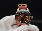 The Vintner SJT Original wristwatch with a gold-tone case, brown dial, blue and red bezel, and red fabric strap, held in a white gloved hand against a black background.