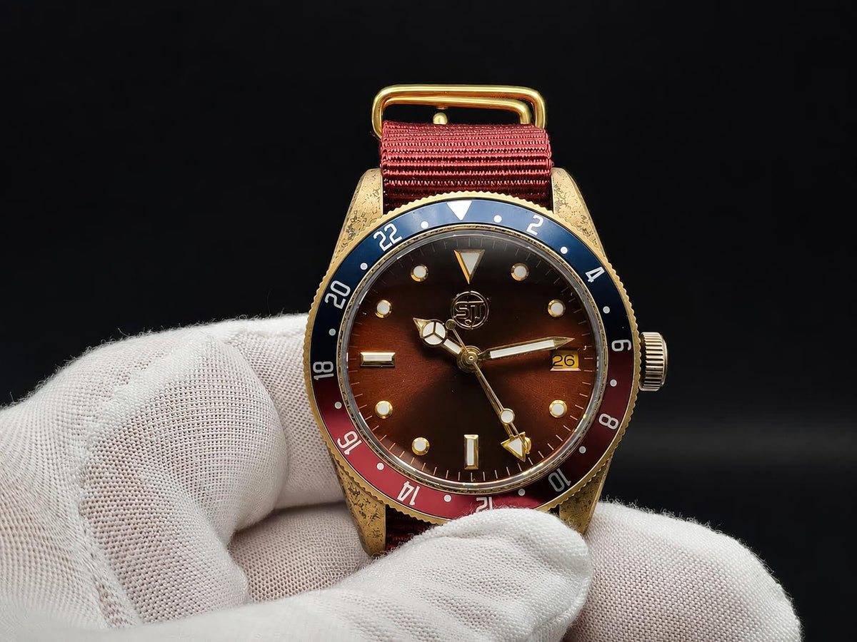 The Vintner SJT Original wristwatch with a gold-tone case, brown dial, blue and red bezel, and red fabric strap, held in a white gloved hand against a black background.