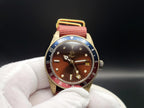 The Vintner SJT Original wristwatch with a brown dial, gold-tone case, red and blue bezel, and red fabric strap, held in a white-gloved hand against a dark background.