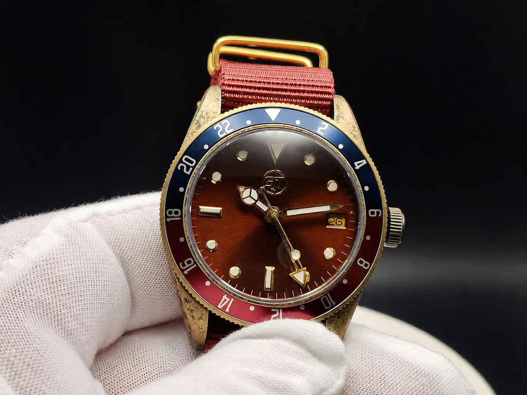 The Vintner SJT Original wristwatch with a gold-tone case, brown dial, blue and red bezel, and red fabric strap, held in a white gloved hand against a black background.