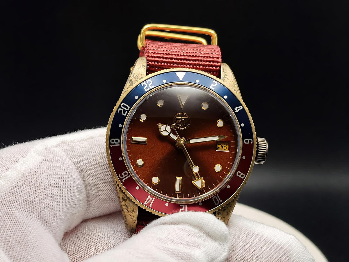 The Vintner SJT Original wristwatch with a gold-tone case, brown dial, blue and red bezel, and red fabric strap, held in a white gloved hand against a black background.