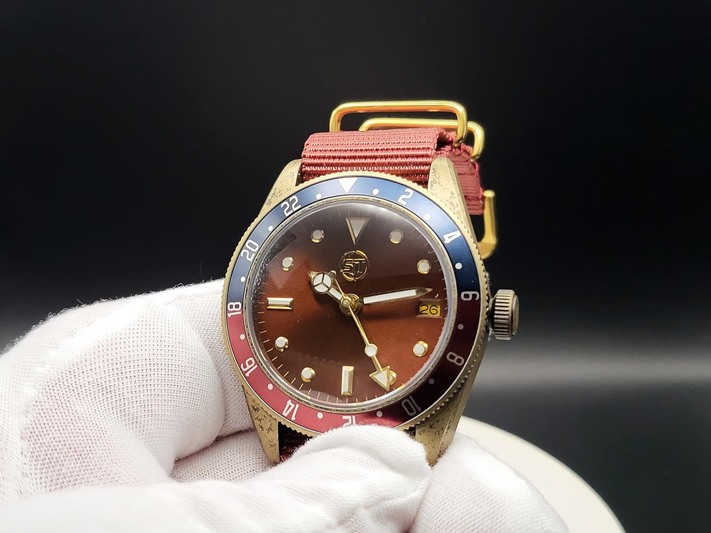 The Vintner SJT Original wristwatch with a gold-toned case, brown dial, red and blue bezel, and red fabric strap, held in a white gloved hand against a dark background.