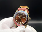 The Vintner SJT Original wristwatch with a gold-toned case, brown dial, red and blue bezel, and red fabric strap, held in a white gloved hand against a dark background.