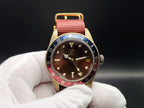 The Vintner SJT Original wristwatch with a brown dial, gold-tone case, red and blue bezel, and red fabric strap, held by a white gloved hand against a dark background.