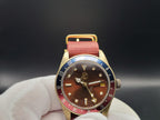 The Vintner SJT Original wristwatch with a round gold-tone case, brown dial, and a red-and-blue bezel, held in a white gloved hand against a dark gray background.