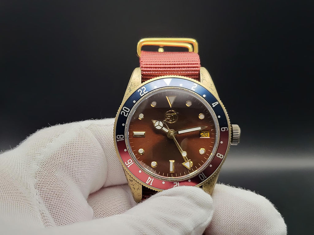 The Vintner SJT Original wristwatch with a gold-tone case, brown dial, and a distinctive red and blue bezel, held by a white gloved hand against a dark background.