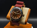 The Vintner SJT Original wristwatch with a brown dial, blue and red bezel, and burgundy fabric strap, displayed on a wooden stand against a dark background.