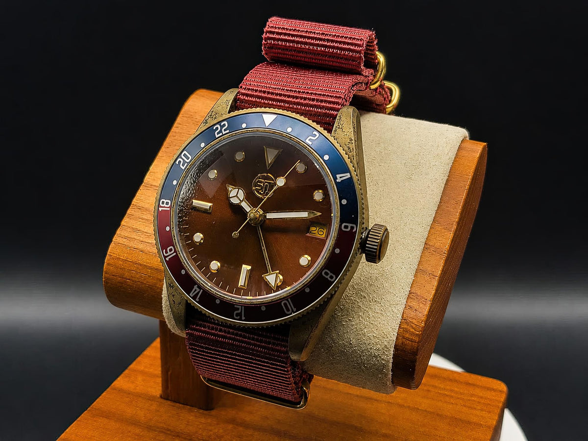The Vintner SJT Original wristwatch with a brown dial, blue and red bezel, and burgundy fabric strap, displayed on a wooden stand against a dark background.