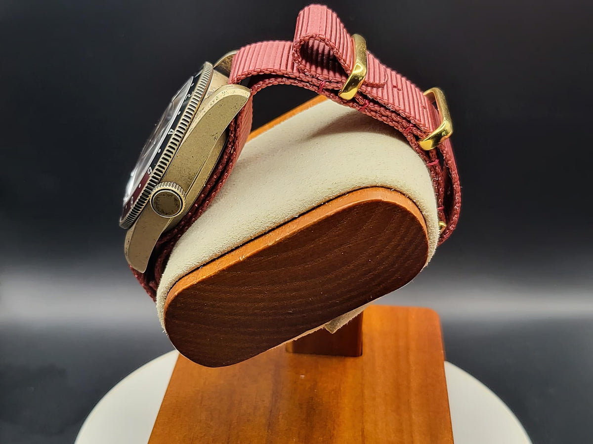 A close-up of The Vintner SJT Original wristwatch with a textured pinkish-red woven strap and gold-tone hardware, displayed on a cream cushion mounted on a wooden stand against a dark background.