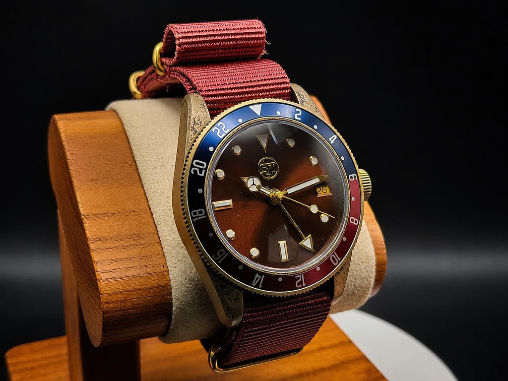 The Vintner SJT Original wristwatch with a brown dial, gold-tone case, and a red-and-blue bezel, displayed on a wooden stand against a dark background.