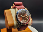 The Vintner SJT Original wristwatch with a round brown dial, a blue and red bezel, and a red fabric strap, displayed on a wooden stand against a dark background.