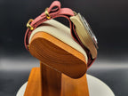 The Vintner SJT Original wristwatch with a dusty rose pink woven strap and gold-tone hardware, displayed on a wooden stand with a cream cushion against a dark background.