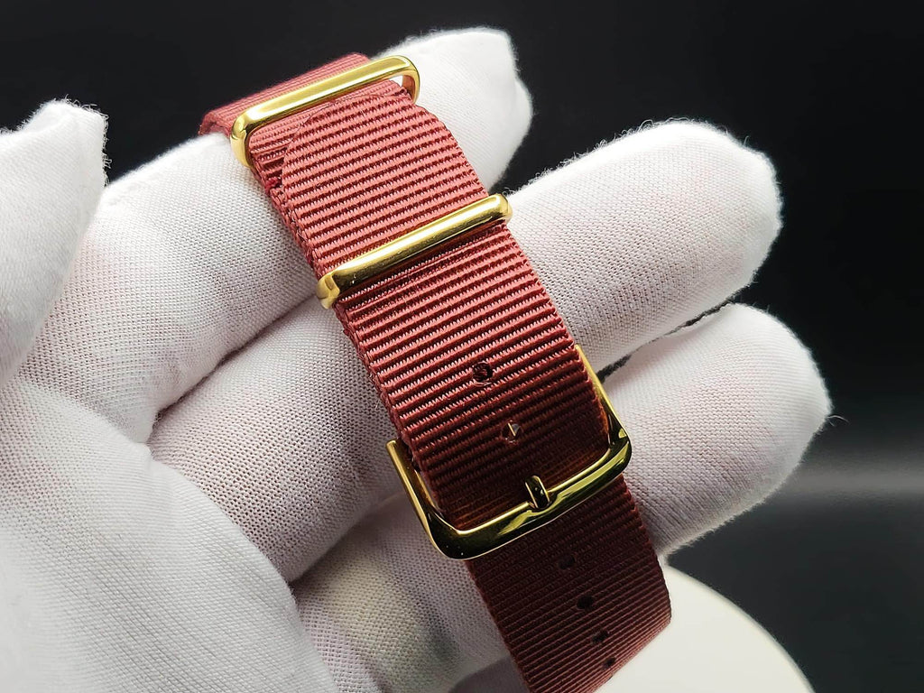 A close-up of a watch strap in textured rose red nylon with polished gold-tone hardware, for The Vintner SJT Original wristwatch, held by a white gloved hand against a dark background.