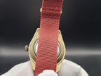 The Vintner SJT Original wristwatch with a red woven nylon strap and a gold-toned metal case, held by a white gloved hand against a dark background.