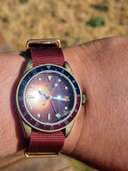 The Vintner SJT Original wristwatch with a bronze-toned case, a dark blue and red bezel, and a burgundy fabric strap, shown worn on a person's wrist outdoors.