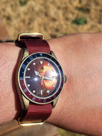 The Vintner SJT Original wristwatch with a red fabric strap, gold-tone case, and a blue and red rotating bezel, shown worn on a person's wrist outdoors.