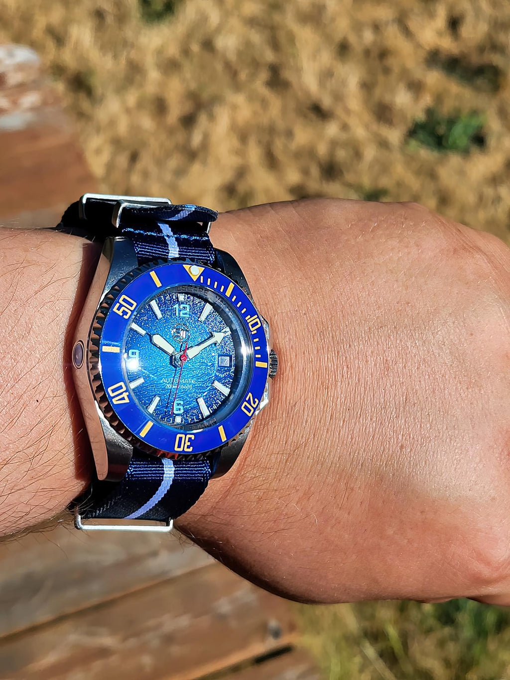 The Bigger Boat SJT Original wristwatch with a blue sunburst dial and a blue-and-black patterned strap, shown worn on a person's wrist against an outdoor blurred background.