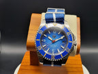 The Bigger Boat SJT Original wristwatch with a blue textured dial, blue bezel, and blue-and-white striped fabric strap, displayed on a wooden stand against a dark background.