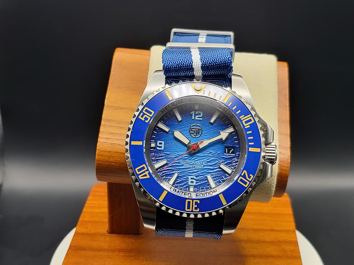 The Bigger Boat SJT Original wristwatch with a blue textured dial, blue bezel, and blue-and-white striped fabric strap, displayed on a wooden stand against a dark background.