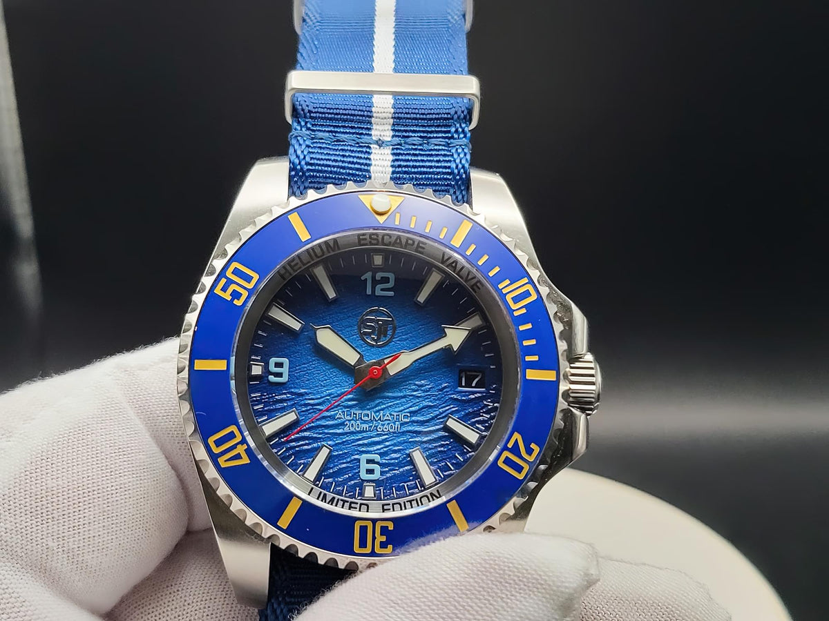 A close-up of The Bigger Boat SJT Original wristwatch with a blue dial, blue bezel, and blue striped fabric strap, held in a white-gloved hand against a dark background.