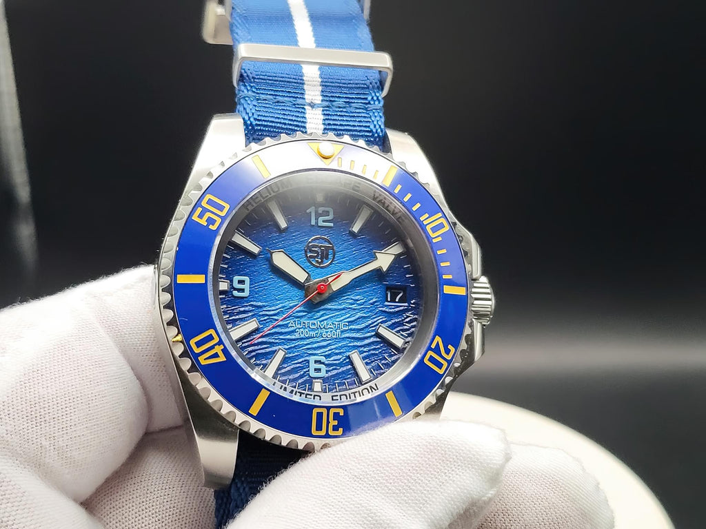 A close-up of The Bigger Boat SJT Original wristwatch with a blue dial, blue bezel, and blue fabric strap with white stripes, held by a white-gloved hand against a dark background.