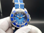 A close-up of The Bigger Boat SJT Original wristwatch with a blue dial, blue bezel, and blue fabric strap with white stripes, held by a white-gloved hand against a dark background.