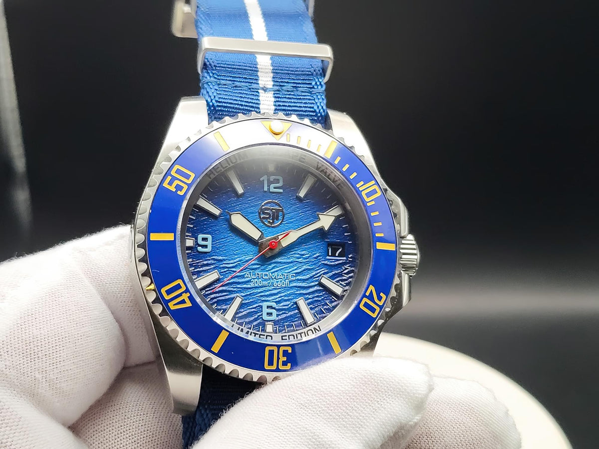 A close-up of The Bigger Boat SJT Original wristwatch with a blue dial, blue bezel, and blue fabric strap with white stripes, held by a white-gloved hand against a dark background.