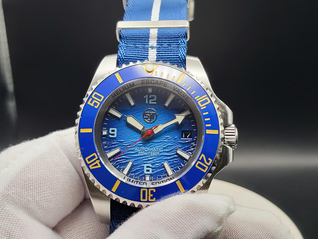 The Bigger Boat SJT Original wristwatch with a blue dial, blue bezel, and blue fabric strap with white stripes, held by a white gloved hand against a dark background.