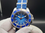 The Bigger Boat SJT Original wristwatch with a blue dial, blue bezel, and blue fabric strap with white stripes, held by a white gloved hand against a dark background.