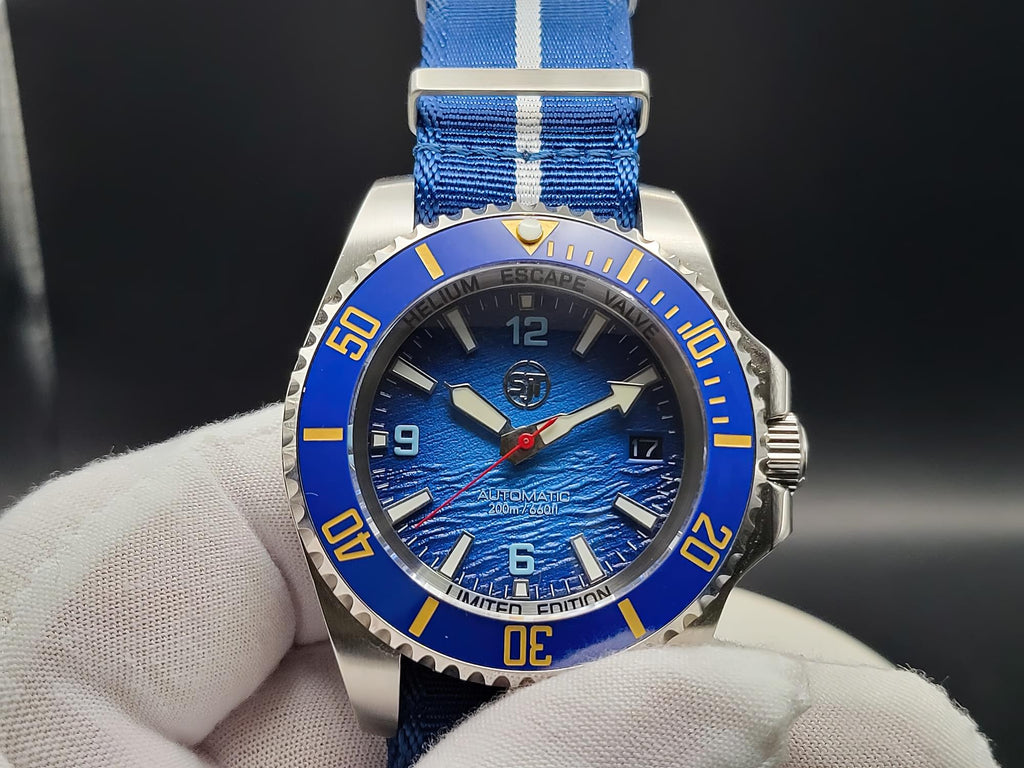 A close-up of The Bigger Boat SJT Original wristwatch with a blue dial, blue bezel, and blue fabric strap with white stripes, held by a white gloved hand against a dark background.