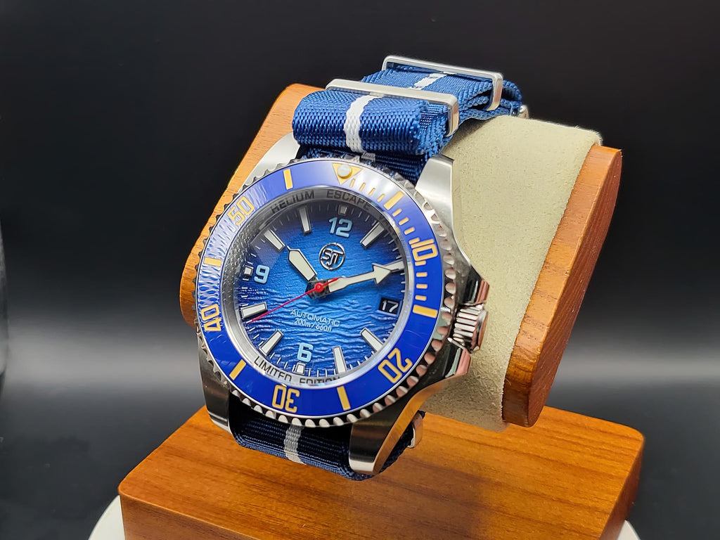 The Bigger Boat SJT Original wristwatch with a blue dial, blue bezel, and blue-and-white striped fabric strap, displayed on a wooden stand against a dark studio background.