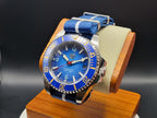 The Bigger Boat SJT Original wristwatch with a blue dial, blue bezel, and blue-and-white striped fabric strap, displayed on a wooden stand against a dark studio background.