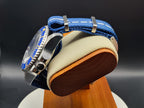 A close-up of The Bigger Boat SJT Original wristwatch with a blue fabric strap and blue bezel detail, displayed on a beige cushion mounted on a wooden stand against a dark background.