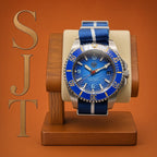 The Bigger Boat SJT Original wristwatch with a blue dial and blue-and-white striped fabric strap, displayed on a wooden stand against an orange background. Large gold letters 'SJT' appear vertically along the left side.