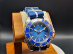The Bigger Boat SJT Original wristwatch with a blue dial, blue and white striped fabric strap, and a silver-tone stainless steel case, displayed on a wooden stand against a dark background.
