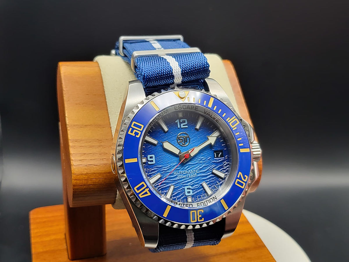 The Bigger Boat SJT Original wristwatch with a blue dial, blue and white striped fabric strap, and a silver-tone stainless steel case, displayed on a wooden stand against a dark background.