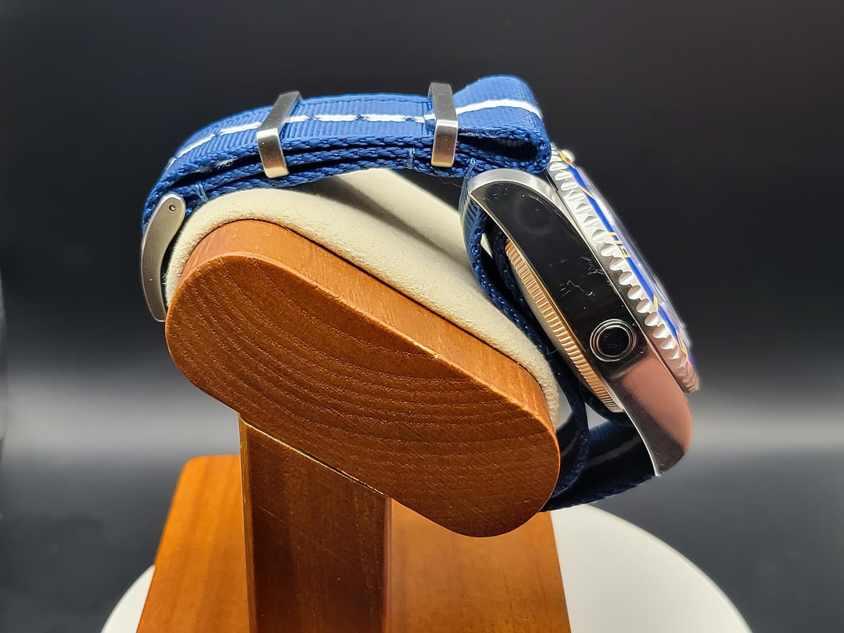A close-up side view of The Bigger Boat SJT Original wristwatch with a blue fabric strap and silver-toned case, displayed on a wooden stand against a dark background.