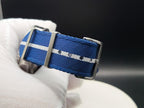A close-up of a blue nylon on The Bigger Boat SJT Original wristwatch strap with a white stripe and silver metal buckle, held by a white gloved hand.