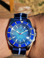 A close-up shot of a wrist wearing The Bigger Boat SJT Original wristwatch with a blue textured dial, a blue and yellow bezel, and a blue striped nylon strap.