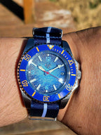 A close-up shot of a wrist wearing The Bigger Boat SJT Original wristwatch with a textured blue dial, a blue bezel with yellow markings, and a blue striped fabric strap.