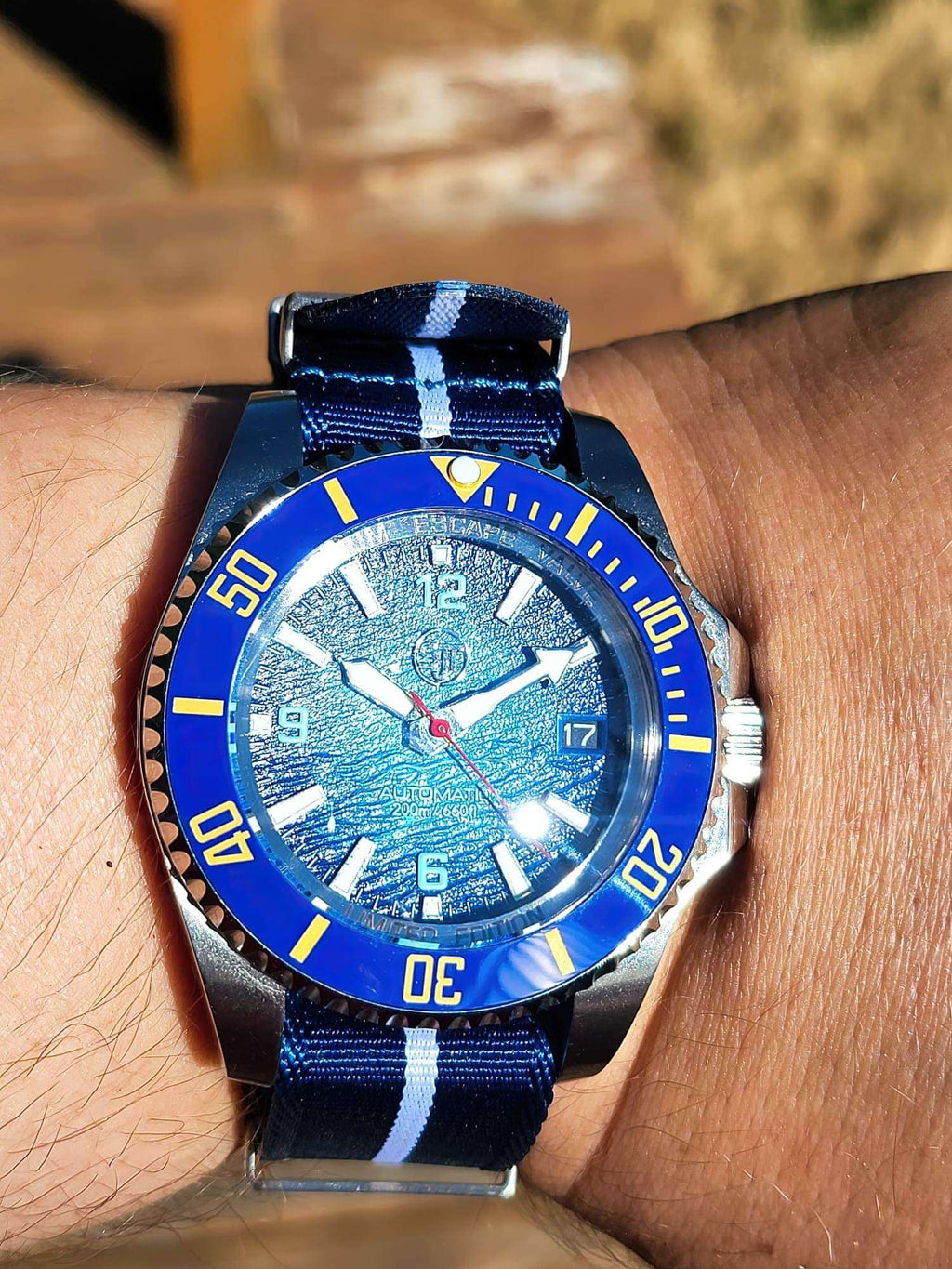 The Bigger Boat SJT Original wristwatch with a blue dial and blue bezel, worn on a person's wrist. The watch has a blue and white striped fabric strap, a silver-toned case, and a blue bezel with yellow numerals.