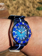 A close-up shot of a wrist wearing The Bigger Boat SJT Original wristwatch with a blue and white striped fabric strap, set against a blurred outdoor background.