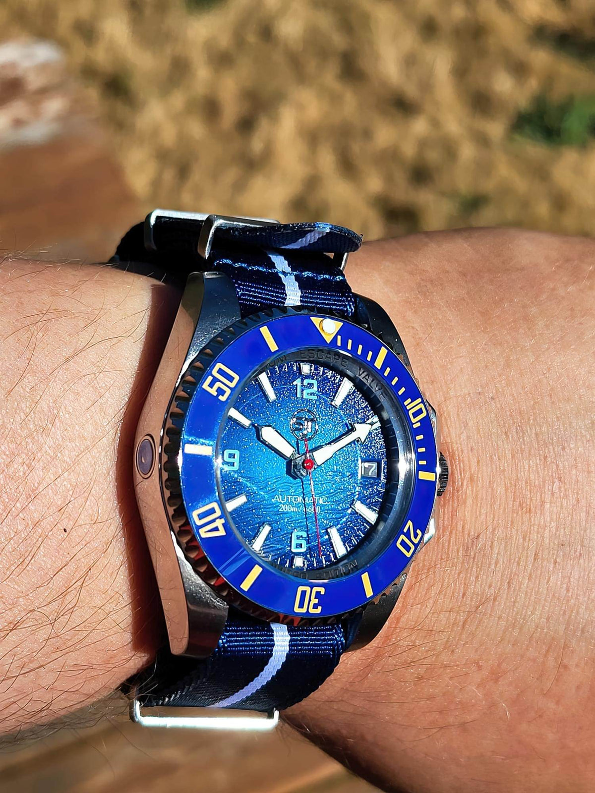 A close-up shot of a wrist wearing The Bigger Boat SJT Original wristwatch with a blue and white striped fabric strap, set against a blurred outdoor background.