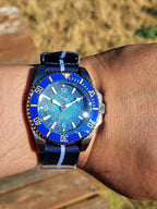 The Bigger Boat SJT Original wristwatch with a blue dial and blue bezel, worn on a person's wrist outdoors. The watch has a blue and white striped fabric strap and a silver-toned case.
