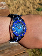 The Bigger Boat SJT Original wristwatch with a blue dial and blue bezel, worn on a person's wrist outdoors. The watch has a dark blue fabric strap with white stripes and a silver-toned metal case.