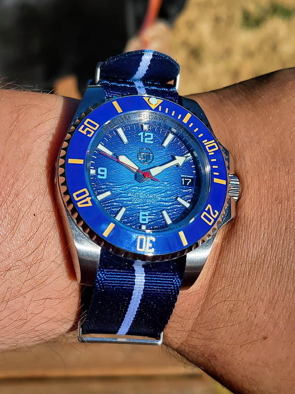 A close-up of a wrist wearing The Bigger Boat SJT Original wristwatch with a blue and white striped fabric strap, photographed outdoors on a person's arm.