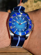 A close-up of a wrist wearing The Bigger Boat SJT Original wristwatch with a blue dial, blue bezel, and blue-and-white striped fabric strap, photographed outdoors on a person's arm.